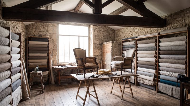 Intérieur de l’atelier dans un ancien chai : charpente apparente, rouleaux de tissus sur étagères, deux fauteuils en cours de restauration sur tréteaux, grande verrière au nord